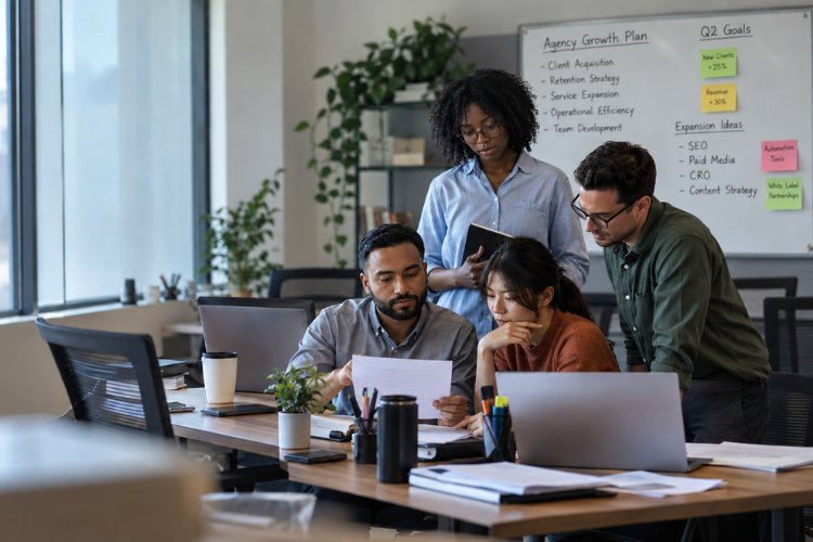 An agency team of four reviewing documents together at a desk of laptops, in front of a whiteboard headed Agency Growth Plan with quarterly goals and expansion ideas