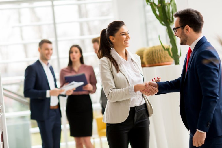 Two professionals shaking hands in a bright office lobby while colleagues talk in the background, illustrating choosing the right partner