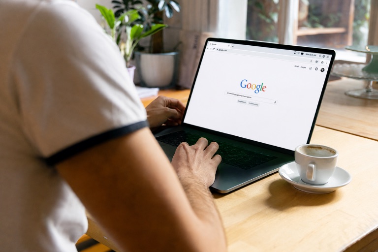 A person at a desk typing a query into the Google homepage on a laptop, beside a cup of coffee