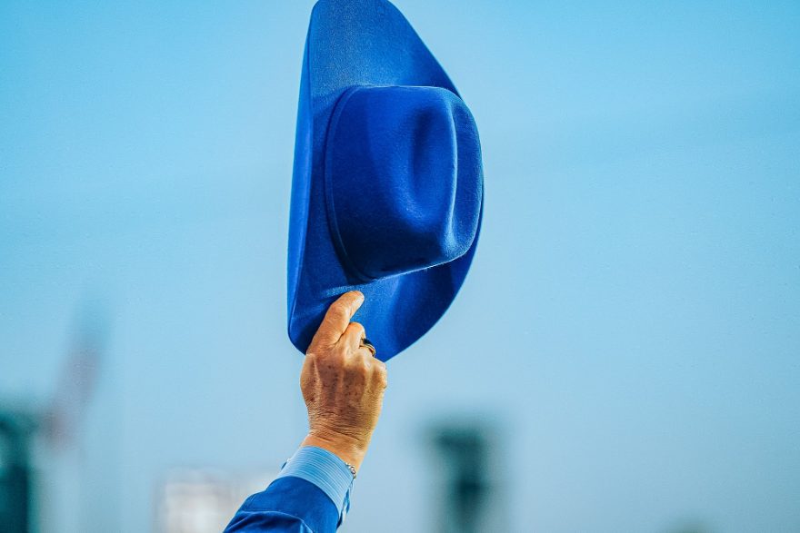A hand raising a blue hat against a clear sky