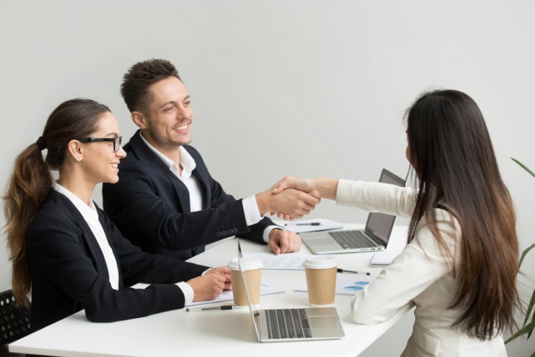 Two agency colleagues greeting a client with a handshake across a desk of laptops, keeping the client relationship consistent and personal