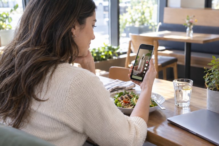 A woman seated at a cafe table viewing content on her smartphone, with a salad, a glass of water and a laptop nearby, representing a customer moving between moments and devices